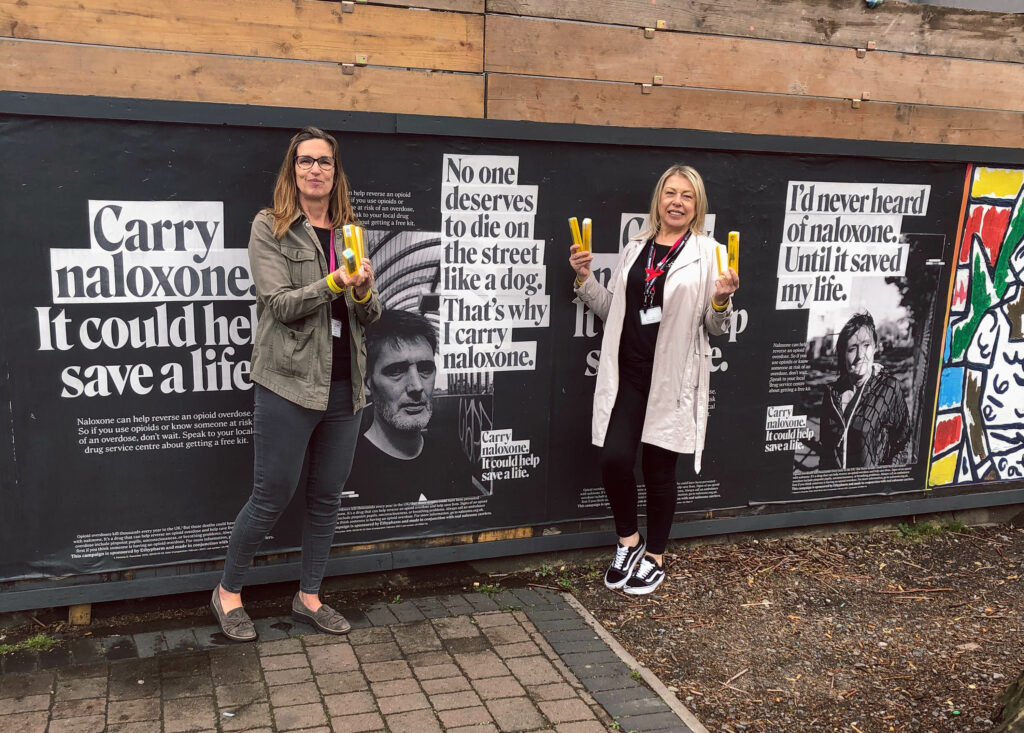 Two BDP workers holding naloxone in front of naloxone poster campaign